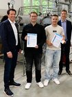 Fünf Männer stehen in einer Versuchshalle vor technischer Ausstattung; zwei junge Männer halten Urkunden zum Master-Förderprogramm im Mauerwerksbau.
